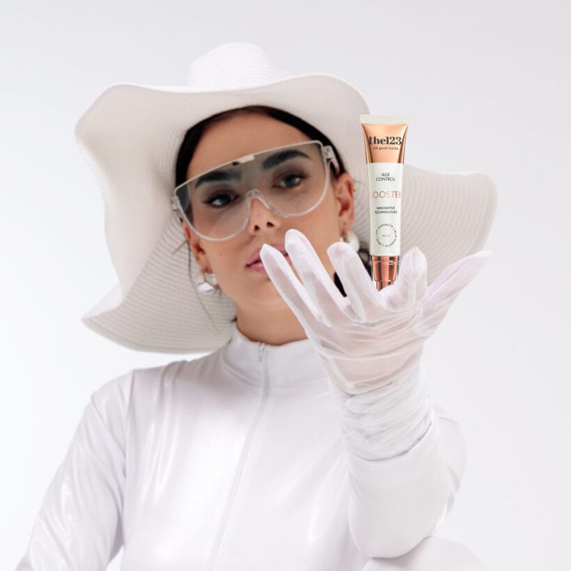 אישה צעירה נאה בלבוש לבן עתידני, מרכיבה משקפיים פוטורוסטיות וחובשת כובע לבן רחב שוליים, כאשר היא מחזיקה בידה שפורפרת של בוסטר אנטי אייגינג 123