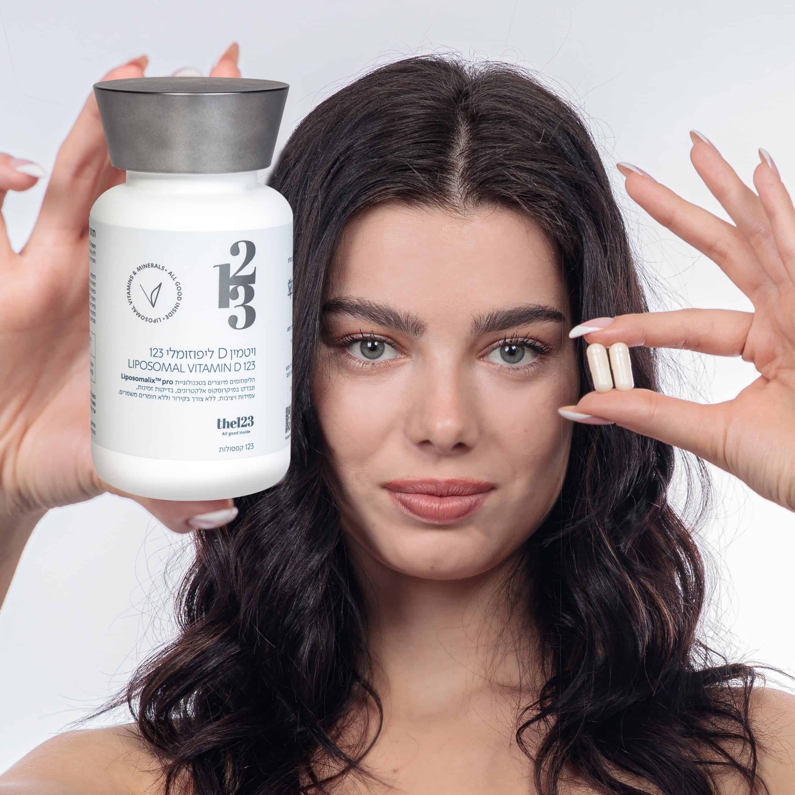אישה צעירה נאה מחזיקה בידה בקבוק של ויטמין די ליפוזומלי 123 ובידה השנייה - 2 קפסולות של תוסף
