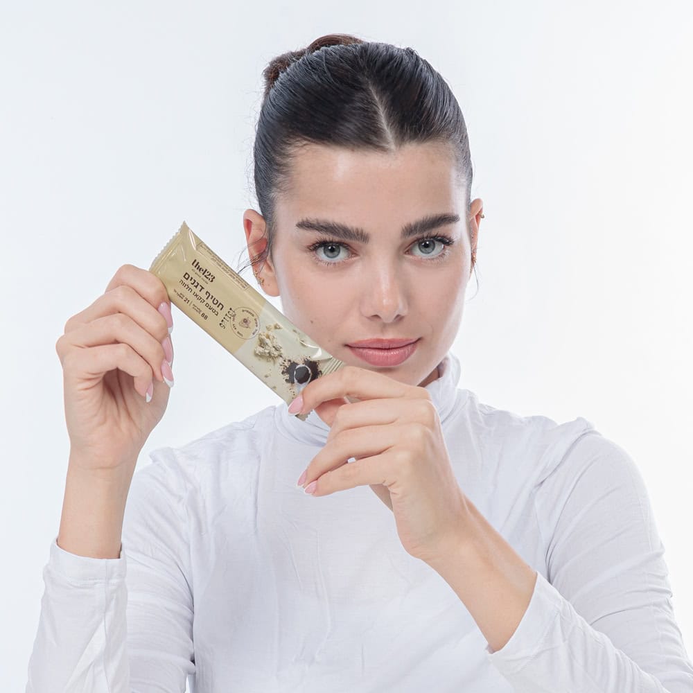 אישה צעירה נאה מחייכת מחזיקה בידיה חטיף דגנים מבית the123 בטעם קקאו חלווה