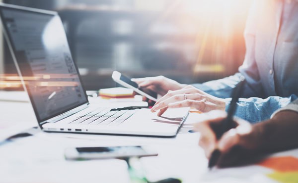 תקריב על ידיהם של שני בני אדם באמצע העבודה במחשב נייד, סמארטפון וכתיבה בעט